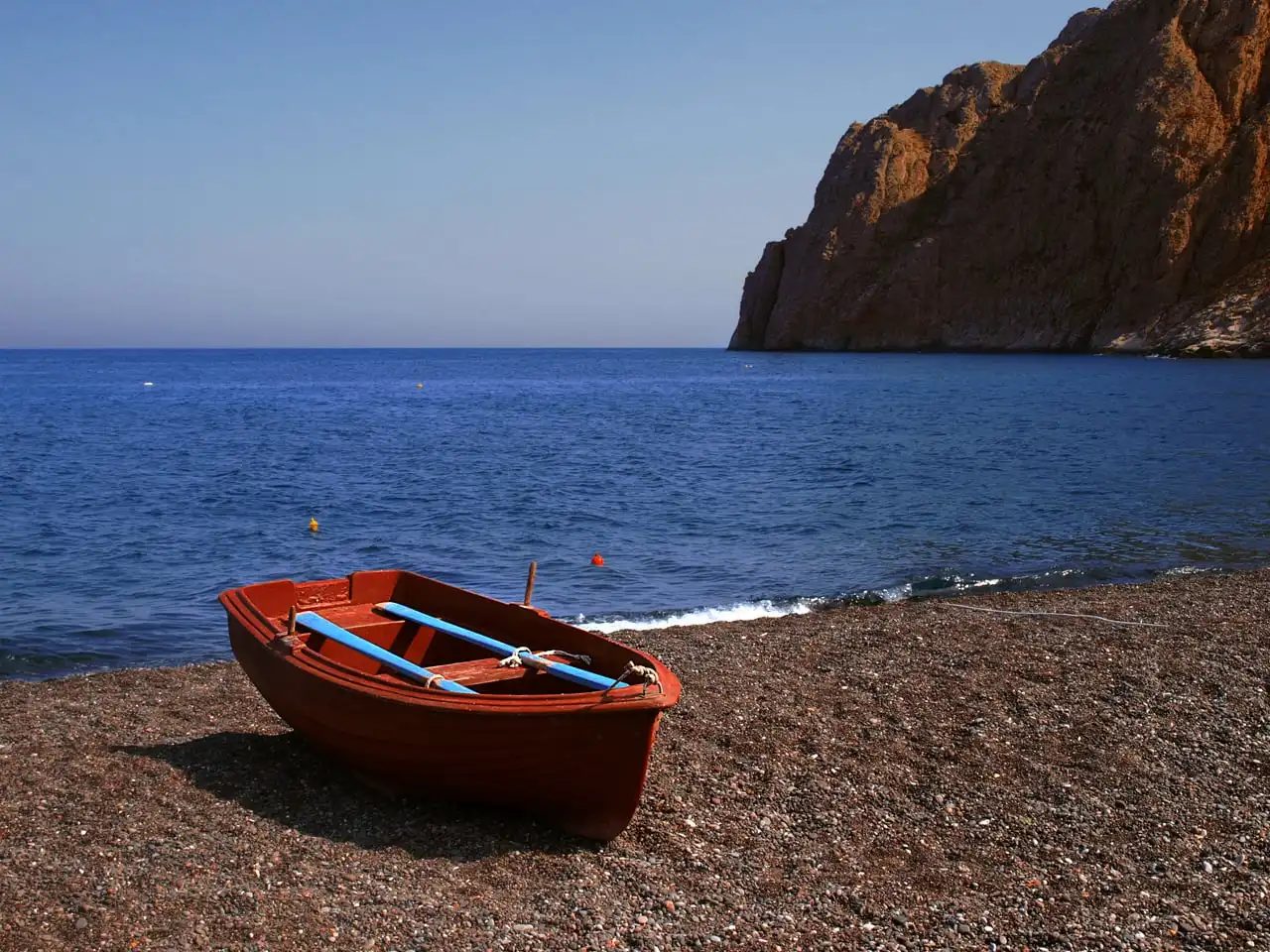 A wooden fishing boat on the black-pebble beach at Kamari, Mesa Vouno rising behind — the flat east-coast shoreline where Gaia works at the waterline