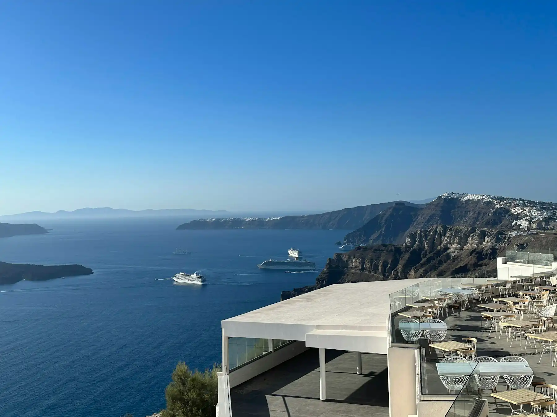 Café tables on a white caldera-edge terrace, cruise ships and the crater rim far below