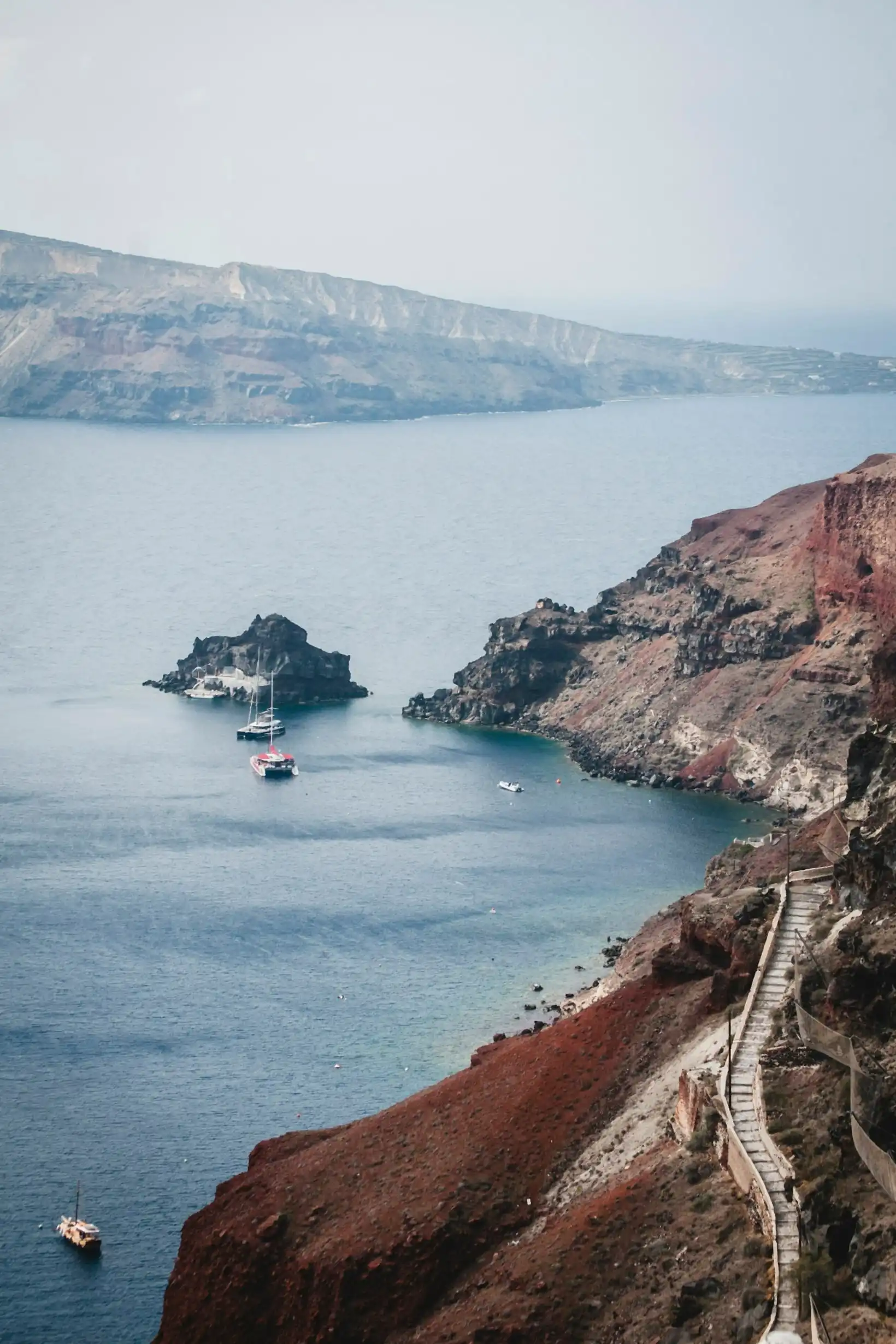 Red volcanic cliffs above Ammoudi Bay, catamarans anchored in the clear water below the Oia headland