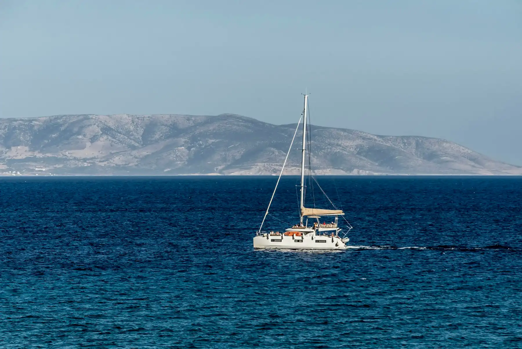 A catamaran motoring across deep blue water below dry Cycladic hills