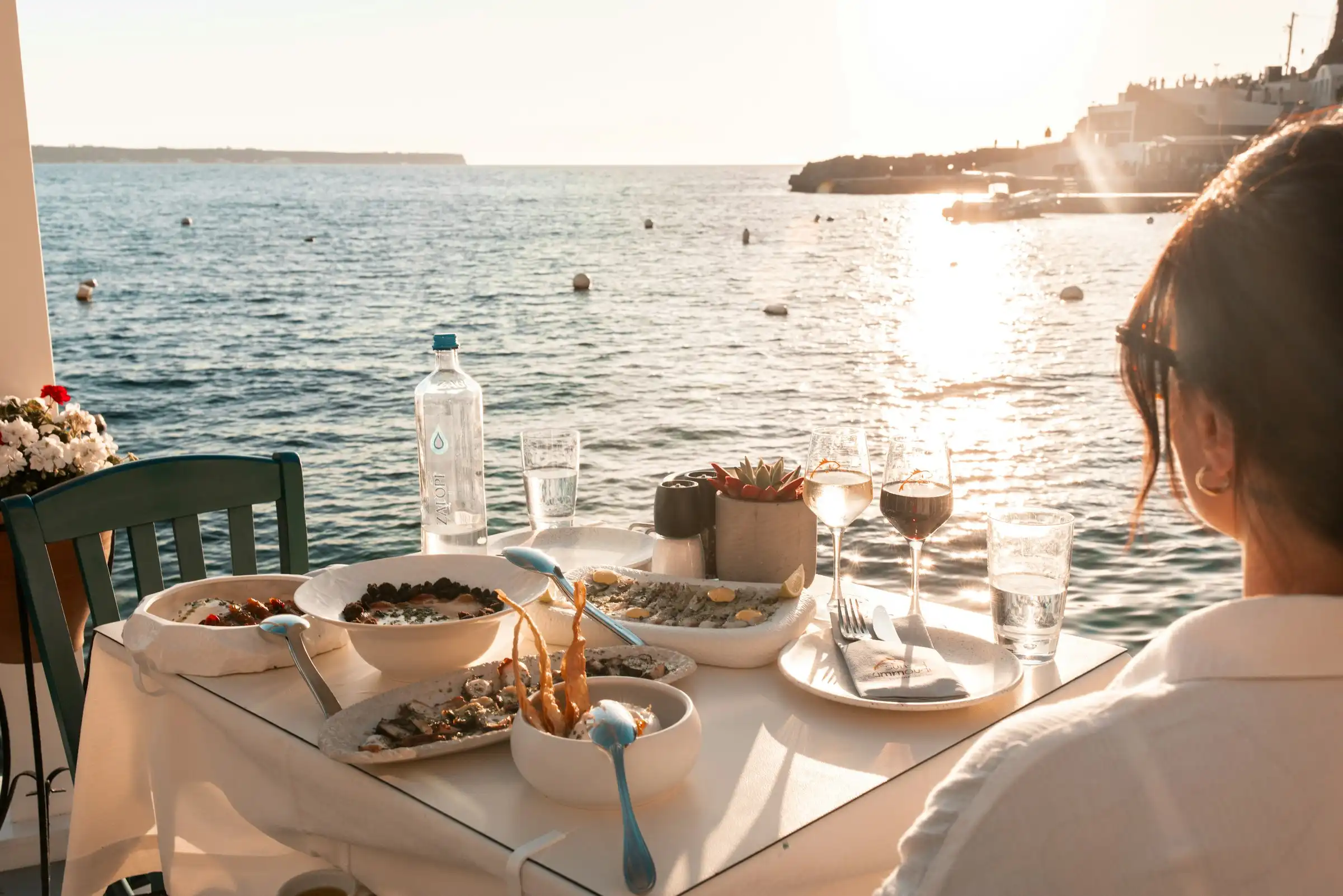 A waterside taverna table at golden hour — white and red wine poured, meze plates open, the Aegean glittering behind