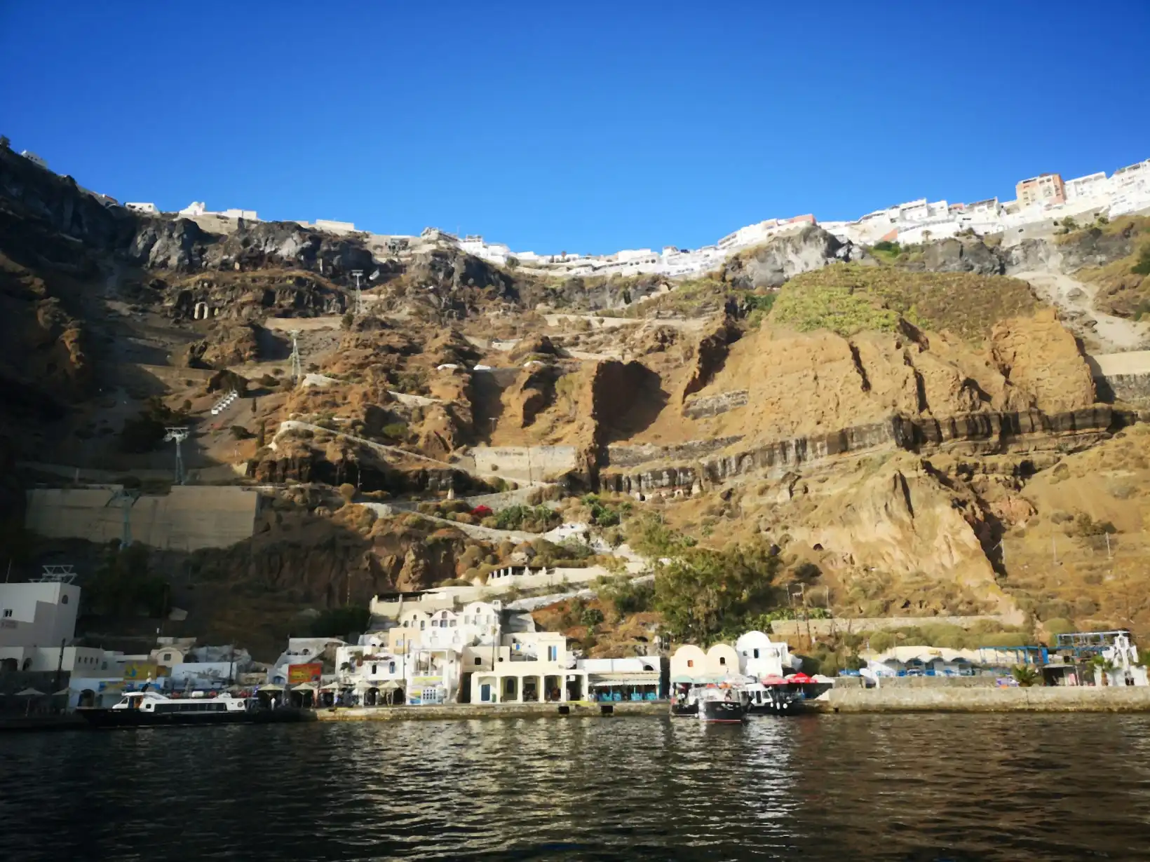 The layered volcanic cliff of Fira rising from the old port, the zig-zag mule path and cable car climbing to the white town on the rim