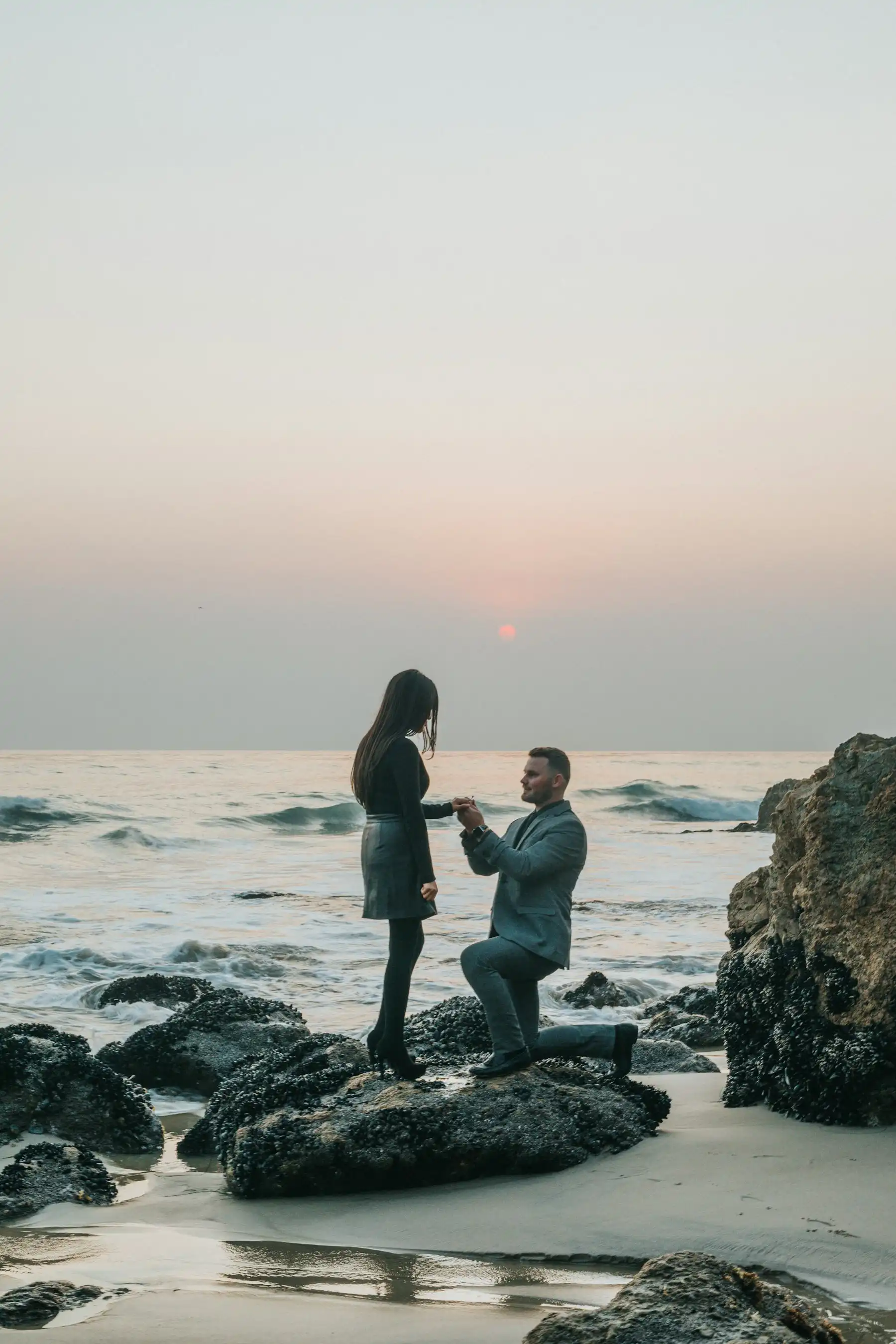 A proposal on a rocky shore at dusk — one partner kneeling, the sun low on the water