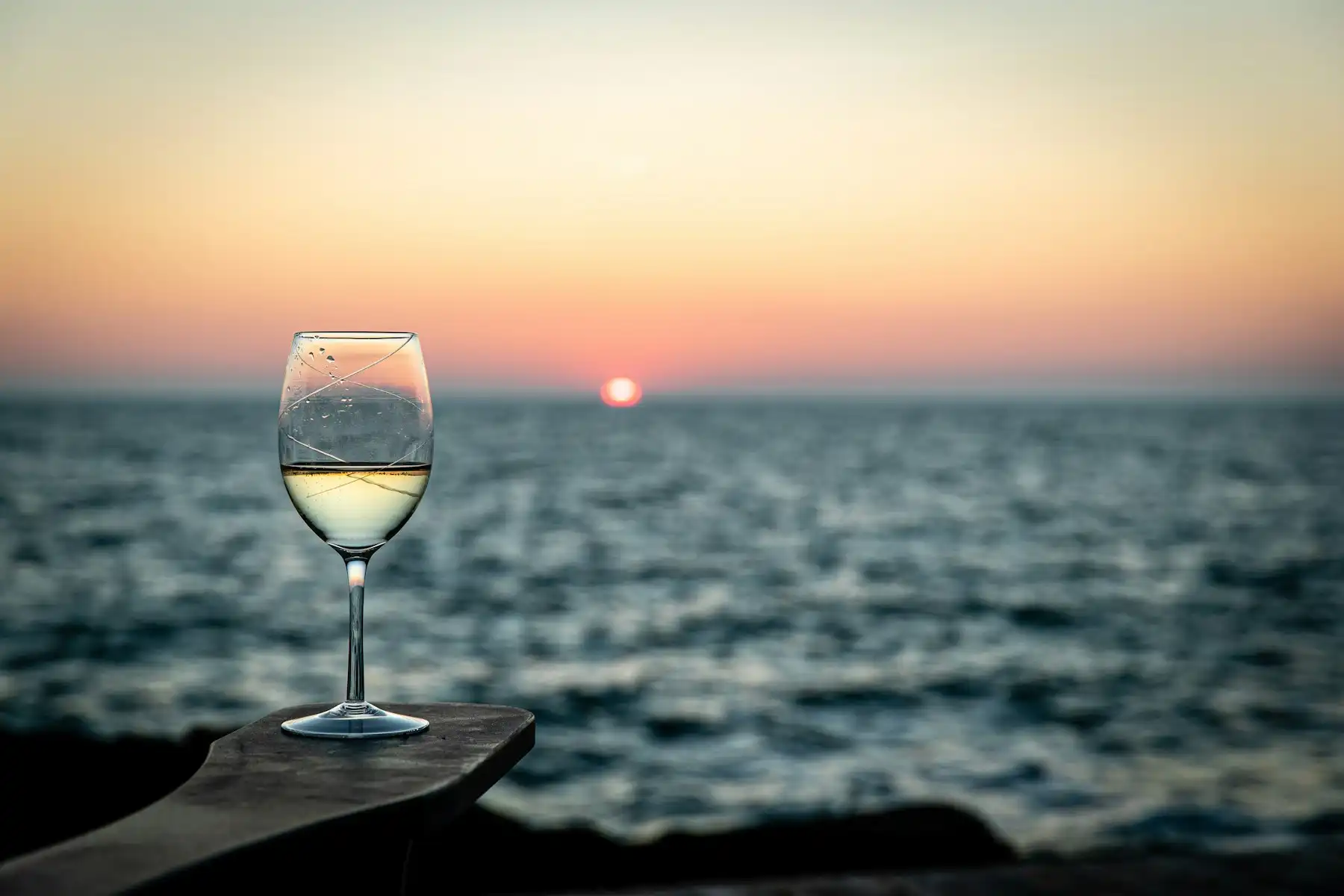 A glass of white wine on a stone ledge as the sun touches the horizon over the sea