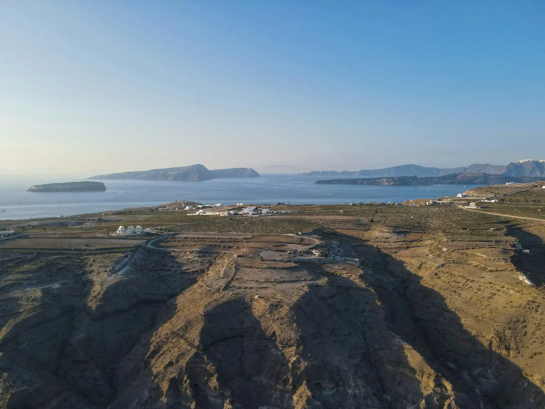 The volcanic plain of southern Santorini from above: ash-grey ravines, scattered farmhouses, the caldera islets offshore