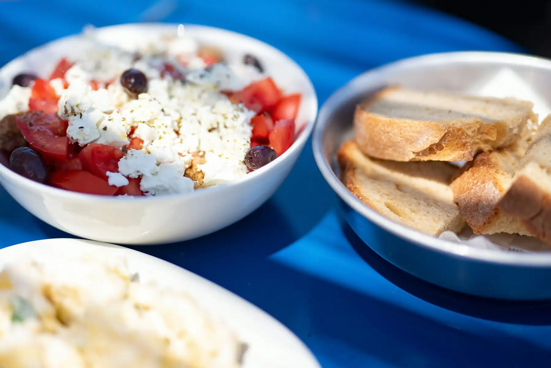 Greek salad with feta and olives beside a dish of barley rusks on a blue taverna table in hard summer sun