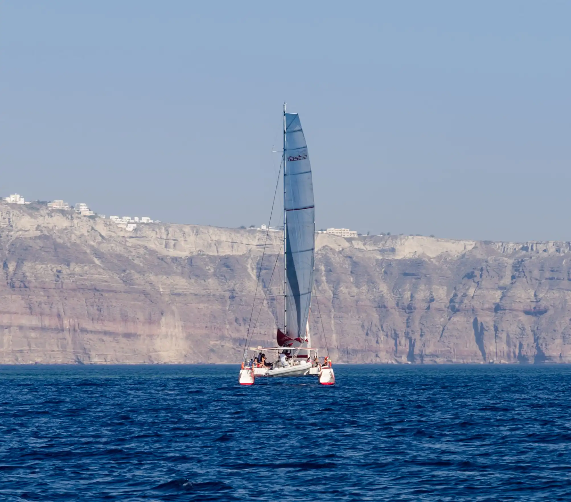 A catamaran under sail beneath the layered caldera cliffs of Santorini, guests out on deck