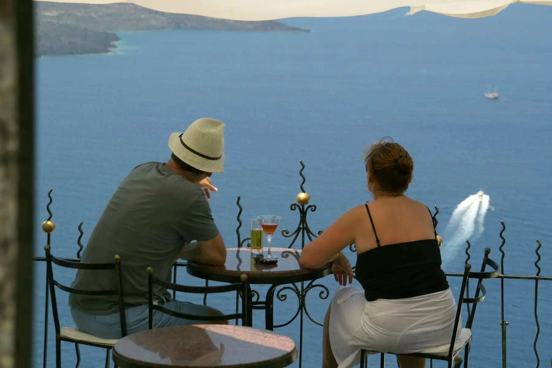 Two travelers at a caldera-edge bar table, drinks between them and the sea far below