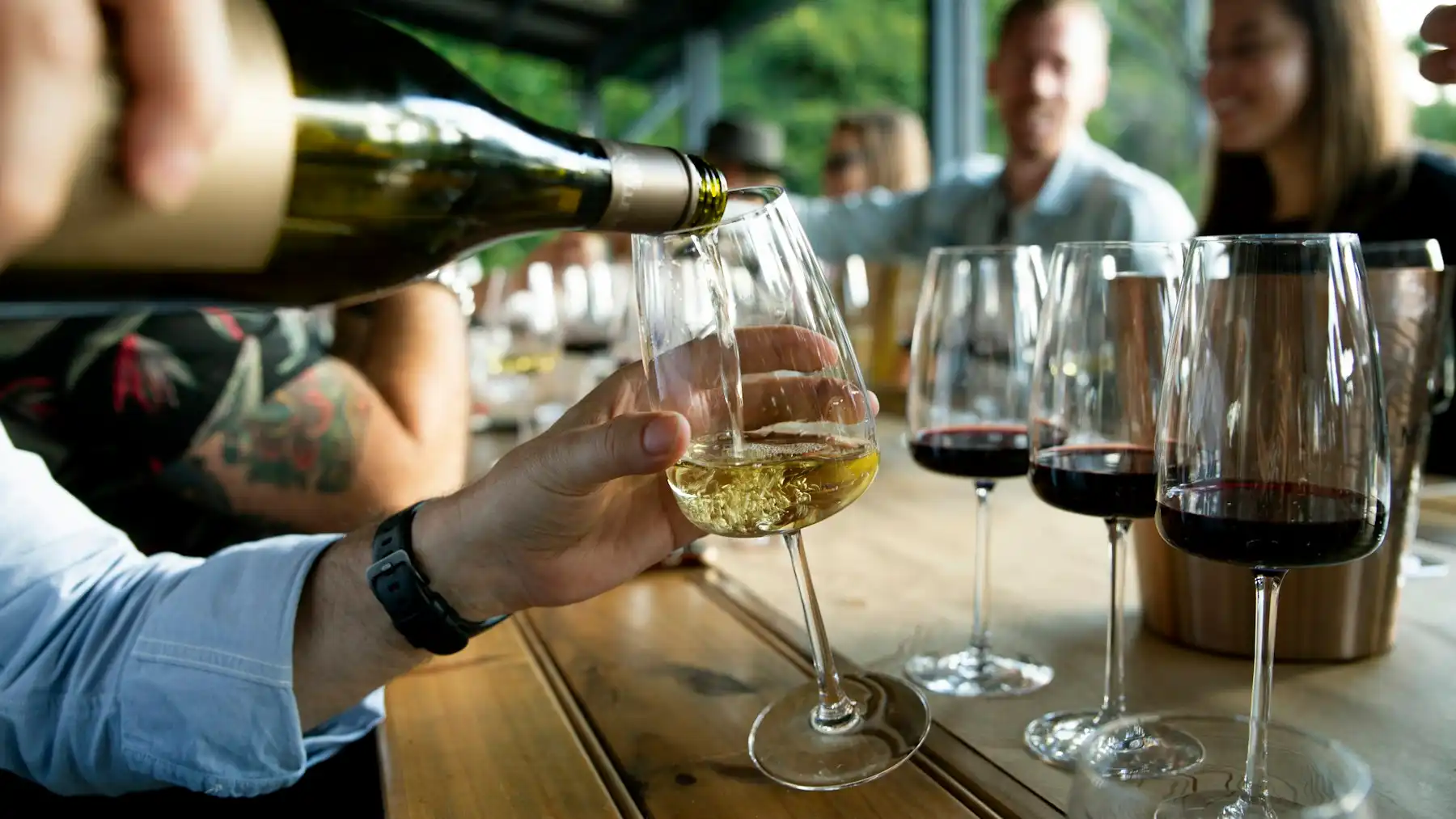 A host pouring white wine for a table of guests, red-wine glasses waiting their turn