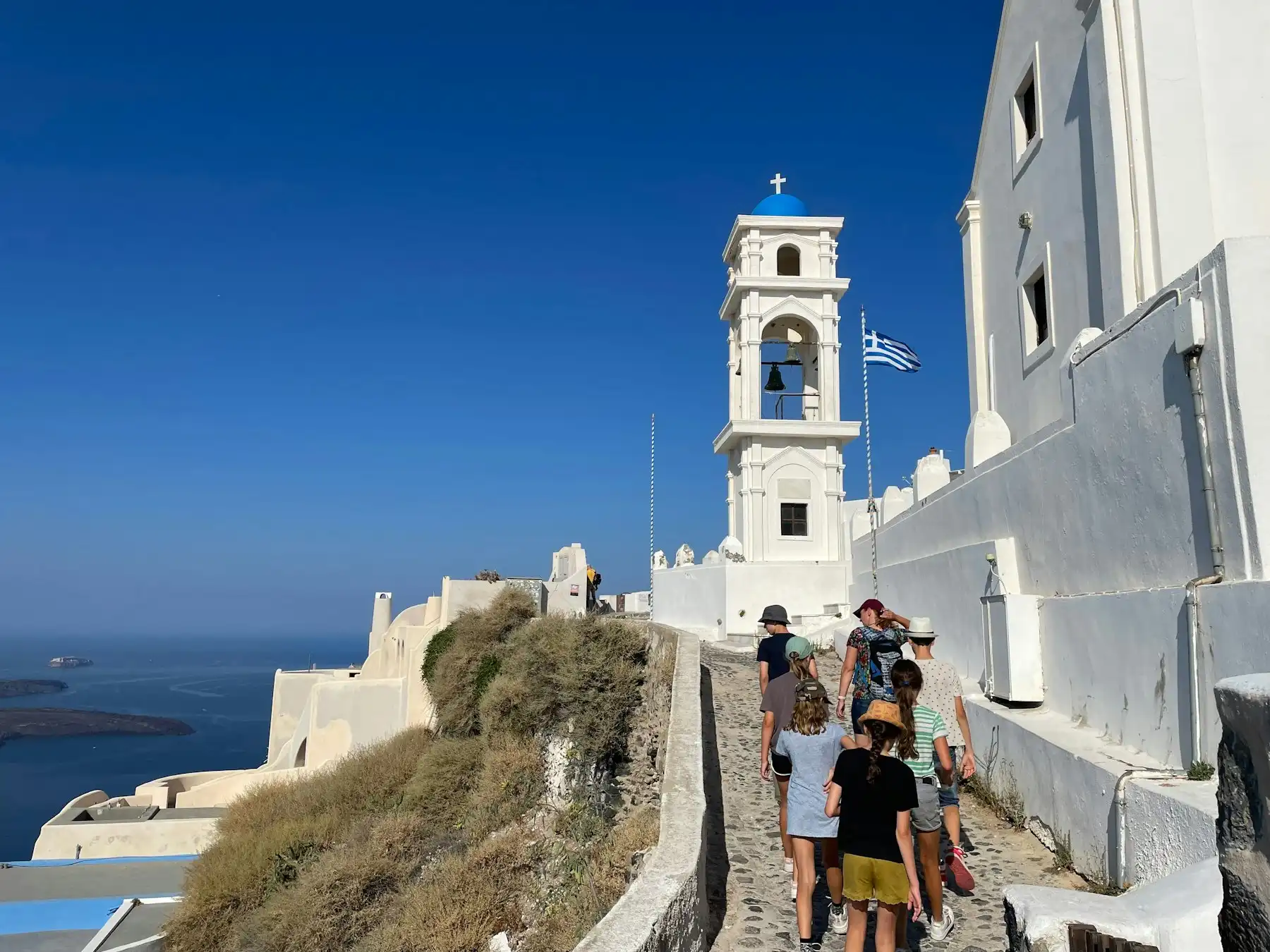 Travelers in sun hats and flat shoes climbing a cobbled path past a bell tower toward the caldera