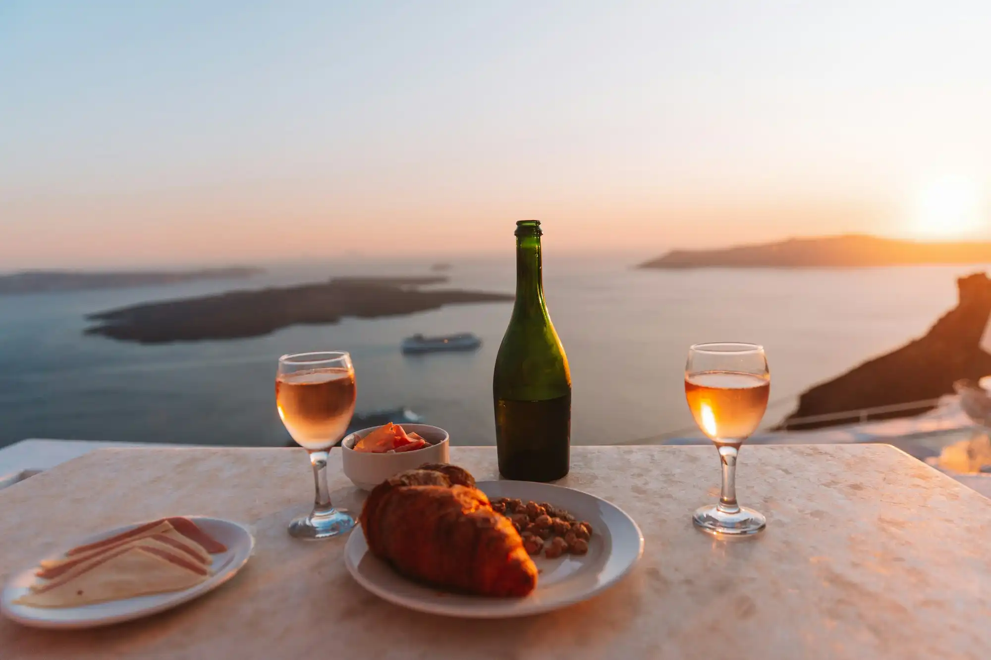 Two glasses of Santorini wine and a bottle on a caldera-edge table at sunset, the volcanic islets and a cruise ship far below