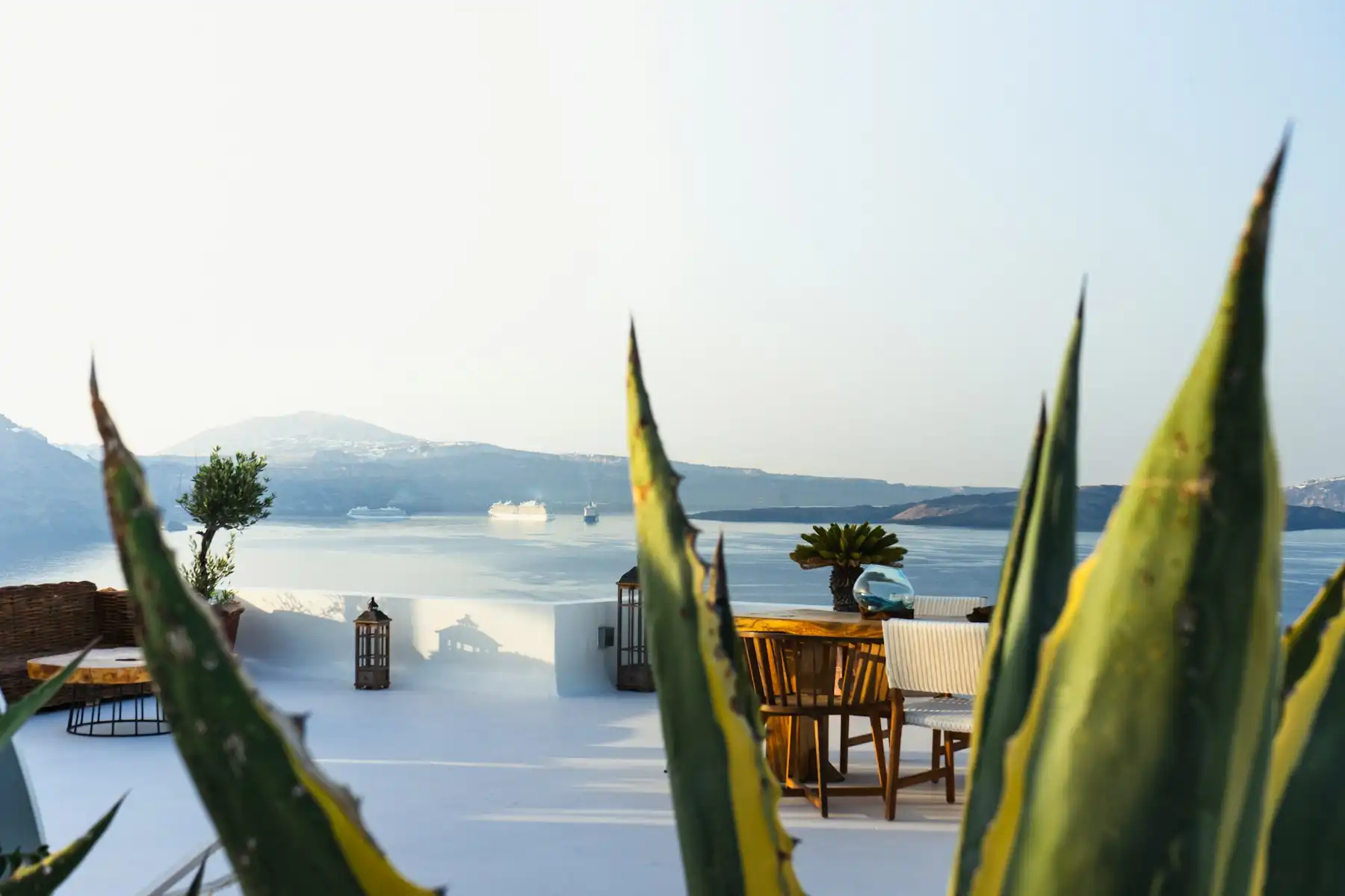 A wooden family table on a rooftop terrace at dusk, agave leaves in the foreground and ships out on the caldera