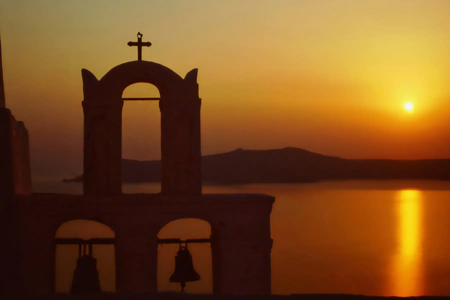 A church bell tower silhouetted against the sun setting behind the caldera rim